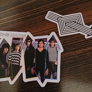 May include: A sticker featuring a band photo of The Cars, a 1970s and 1980s rock band, with the band name in white text. The band members are standing in front of a colorful, abstract background. The sticker is rectangular and has a white border.