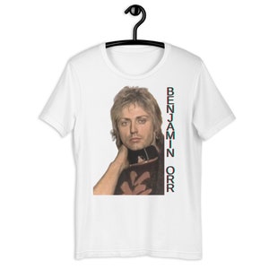 May include: White t-shirt with a photo of a man with blonde hair and a black and white floral shirt. The text "BENJAMIN ORR" is printed vertically on the right side of the shirt.