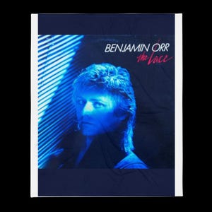 May include: A blue blanket featuring the album cover of Benjamin Orr's "The Lace." The image shows a portrait of a man with blue lighting and the album title in white and pink text.