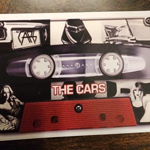 May include: A vintage cassette tape with a red and black design. The tape has the band name "THE CARS" printed on it. The cassette tape has a clear plastic case with a silver metal band.