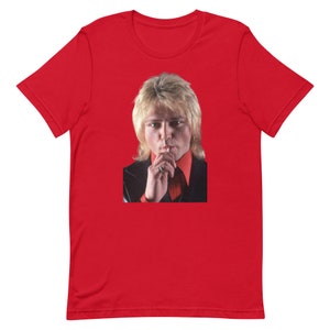 Puede incluir: Una camiseta roja de manga corta con una foto de un hombre con el pelo rubio, que lleva una camisa roja y una chaqueta negra, mirando a la cámara.