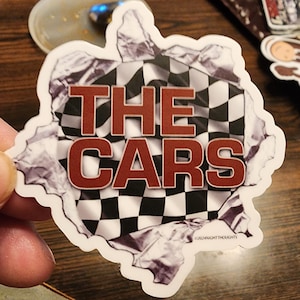 May include: A white sticker with a checkered flag design. The flag is torn to reveal the words "THE CARS" in red letters. The sticker has a copyright notice at the bottom that reads "© Midnight Thoughts".