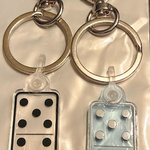May include: Two keychains with heart-shaped clasps and gold-colored key rings. Each keychain has a rectangular, transparent charm with a domino design. One charm has black dots, the other has light blue dots.