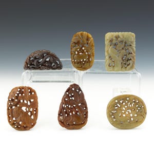 May include: A collection of six carved stone pendants in various shapes and colors. The pendants feature intricate floral and abstract designs. Colors include shades of brown, tan, and cream. The pendants are displayed on a clear acrylic surface.
