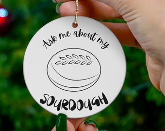 May include: White ceramic Christmas ornament with a black and white illustration of a loaf of sourdough bread. The ornament reads "Ask me about my sourdough".