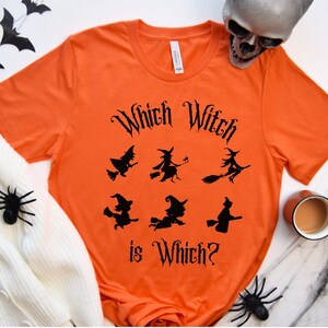 May include: Orange t-shirt with a black graphic that says "Which Witch is Which?" and features five black silhouettes of witches flying on broomsticks.