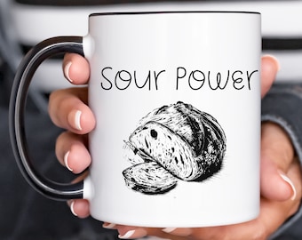 May include: White ceramic mug with a black handle. The mug has a black outline and the text "Sour Power" printed in black. A black and white illustration of a loaf of sourdough bread is below the text.