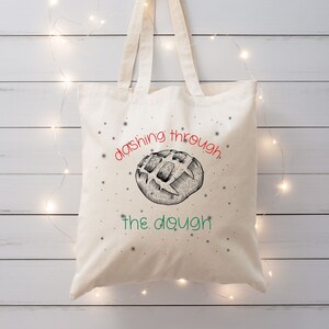 May include: A white canvas tote bag with a black and white illustration of a bread roll and the text "dashing through the dough" in red and green.