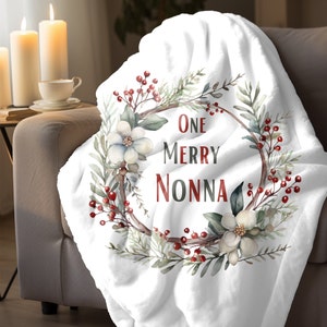 May include: White fleece throw blanket with a watercolor wreath design featuring green leaves, white flowers, and red berries. The wreath has a brown circle in the center with the text "One Merry Nonna" in red and black.