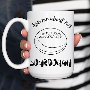 May include: White ceramic mug with a black design that says "Ask me about my sourdough". The design also includes a black and white illustration of a loaf of sourdough bread.