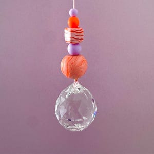 Könnte beinhalten: Ein hängender Sonnenfänger mit einem klaren Kristallkugel am unteren Ende, verbunden mit einer Schnur mit verschiedenen Perlen. Die Perlen umfassen eine große orangefarbene Kugel, eine gestreifte orange-weiß gestreifte Perle und kleinere lila und orangefarbene Perlen.