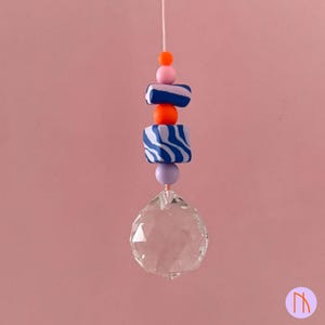 Könnte beinhalten: Ein dekorativer Sonnenfänger mit einer durchsichtigen Kristallkugel am unteren Ende, aufgereiht an einer weißen Schnur. Die Schnur ist mit einer Reihe farbiger Perlen verziert, darunter orangefarbene, rosa, lila und blau-weiß gestreifte quadratische Perlen. Der Hintergrund ist einfarbig rosa.
