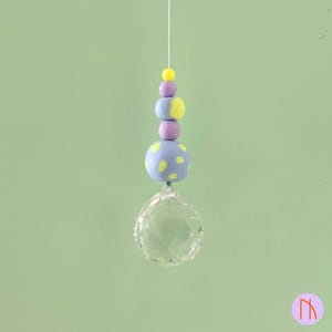 Pode incluir: Uma bola de cristal decorativa suspensa com um fio, adornada com contas coloridas. As contas são em tons de amarelo, roxo e azul, com um padrão de bolinhas. A bola de cristal é transparente e facetada.