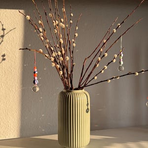 Puede incluir: Un jarrón verde claro con estrías contiene ramas de sauce con pequeños capullos blancos. Adornos de cuentas de colores cuelgan de las ramas. El jarrón está sobre una superficie blanca, con sombras proyectadas en la pared.