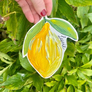 Puede incluir: Ilustración de limón pintada a mano con un cuerpo amarillo brillante y hojas verdes. La palabra "Lucie" está escrita en cursiva en el lado derecho. La obra de arte está sobre un fondo de follaje verde.
