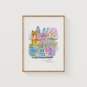 May include: Watercolor print of Cincinnati, Ohio, featuring a bridge, buildings, and a fountain. The artwork is framed in a light brown wooden frame. The print has a colorful, artistic style with the city's name written in cursive.