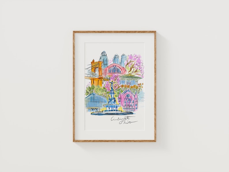 May include: Watercolor print of Cincinnati, Ohio, featuring a bridge, buildings, and a fountain. The artwork is framed in a light brown wooden frame. The print has a colorful, artistic style with the city's name written in cursive.