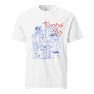 Può includere: T-shirt bianca con grafica blu e rossa raffigurante i monumenti di Kansas City. Il design include le parole "Kansas City" in corsivo rosso e l'insegna Western Auto. Una maglietta casual e confortevole.