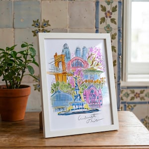 May include: A framed watercolor print of Cincinnati, Ohio, featuring iconic landmarks in vibrant colors. The artwork includes a bridge, buildings, and a fountain, with the city's name handwritten at the bottom. The frame is white.