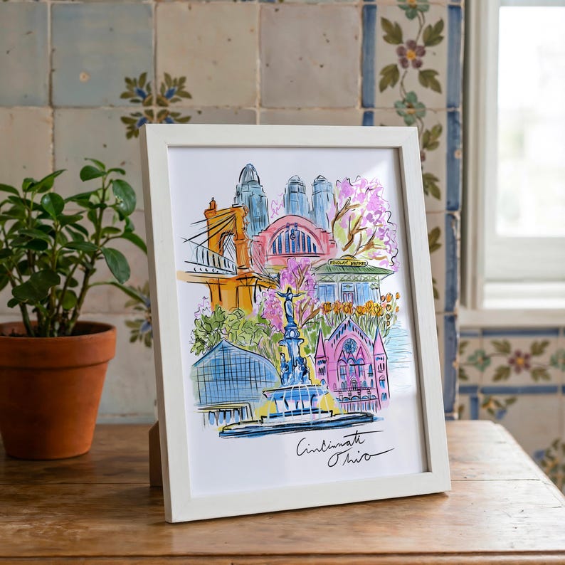 May include: A framed watercolor print of Cincinnati, Ohio, featuring iconic landmarks in vibrant colors. The artwork includes a bridge, buildings, and a fountain, with the city's name handwritten at the bottom. The frame is white.