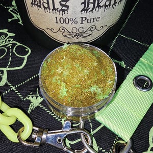 May include: A small clear jar filled with gold glitter and green accents. The jar is attached to a green lanyard with a silver clasp and a silver ring. The jar has a label that reads "Bats Heart 100% Pure".