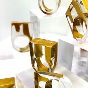 May include: A collection of clear resin rings featuring embedded gold-colored metallic swirls. Each ring has a thick, rectangular profile with a circular opening for the finger, showcasing a transparent body with distinct, flowing gold-toned patterns.