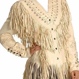 Puede incluir: Una chaqueta de cuero blanca con detalles de flecos y tachuelas plateadas. La chaqueta tiene un cierre de botones en la parte delantera y mangas largas.