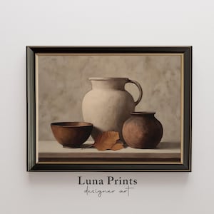 May include: A framed still life print depicting a beige jug with a handle, a small brown vase, and a wooden bowl. A dried leaf sits near the base of the jug. The print has a neutral colour palette.
