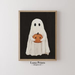 Peut inclure: Un fant&ocirc;me blanc tenant une citrouille orange sculpt&eacute;e sur un fond noir. L'image est encadr&eacute;e dans un cadre en bois brun clair. Le texte "Luna Prints - designer art" est sous le cadre.