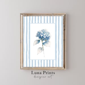 May include: A framed watercolor print featuring a blue hydrangea blossom. The artwork is set against a white background with light blue vertical stripes and a wooden frame. The text "Luna Prints designer art" is at the bottom.
