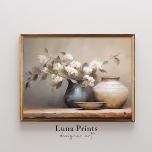 Puede incluir: Pintura enmarcada de naturaleza muerta con un ramo de flores blancas en un jarrón azul oscuro, un jarrón beige y un cuenco pequeño sobre una superficie de madera. La obra de arte tiene una paleta de colores neutros y una estética vintage.