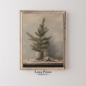 Puede incluir: Una pintura enmarcada de un pequeño pino en una maceta blanca, sentado sobre una superficie de madera con un paño blanco drapeado sobre él. El fondo es de un gris apagado con una textura sutil.