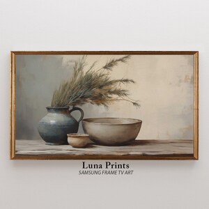 以下が含まれることがあります： 青い陶器のピッチャーに取っ手が付いていて、白い陶器のボウルと小さな茶色の陶器のボウルが置かれた静物画。ピッチャーには乾燥した草が詰められていて、ボウルは空です。絵は金色の額縁に飾られており、下部に「Luna Prints SAMSUNG FRAME TV ART」の文字があります。