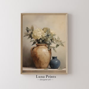 May include: A framed art print depicting a still life. A large, aged brown vase contains white flowers and green leaves. A small blue vase is positioned next to it. The artwork is signed "Luna Prints - designer art -."