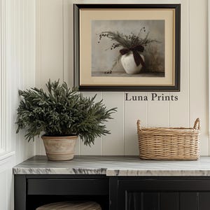 May include: A framed print of a still life featuring a white vase with a burgundy ribbon and dried flowers. The print is hanging on a white wall with a green plant in a terracotta pot and a wicker basket on a black countertop. The text "Luna Prints" is visible below the print.
