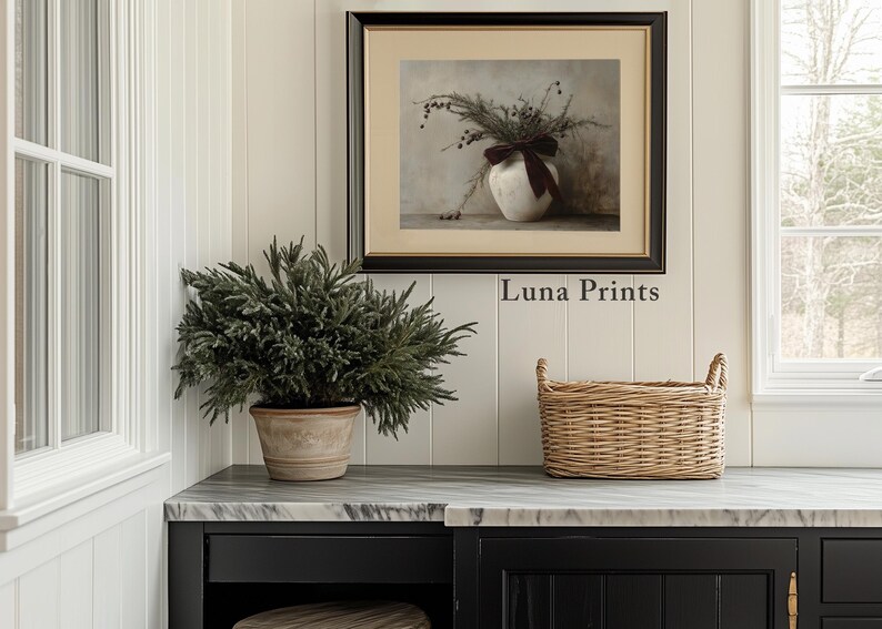 May include: A framed print of a still life featuring a white vase with a burgundy ribbon and dried flowers. The print is hanging on a white wall with a green plant in a terracotta pot and a wicker basket on a black countertop. The text "Luna Prints" is visible below the print.