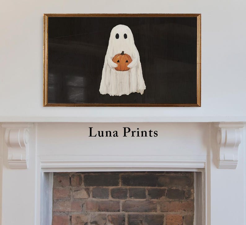 May include: A framed Halloween-themed artwork featuring a white ghost holding an orange pumpkin against a black background. The artwork is in a gold frame and is displayed above a white mantelpiece. The text "Luna Prints" is below the artwork.