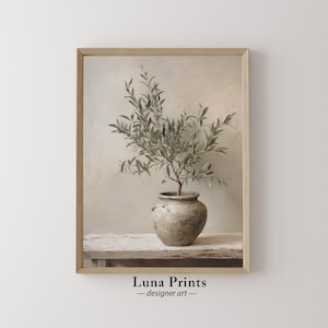 May include: A framed print of a green olive branch in a grey ceramic pot on a white wooden surface. The background is a light beige wall. The print is titled "Luna Prints - designer art".
