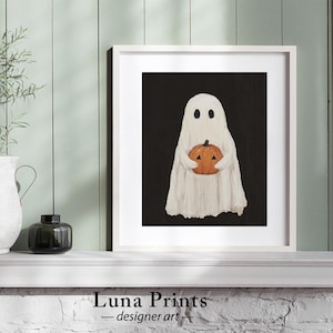 Peut inclure: Un fant&ocirc;me blanc tenant une petite citrouille orange devant un fond noir. Le fant&ocirc;me a deux yeux noirs et une petite bouche. L'image est encadr&eacute;e dans un cadre blanc.