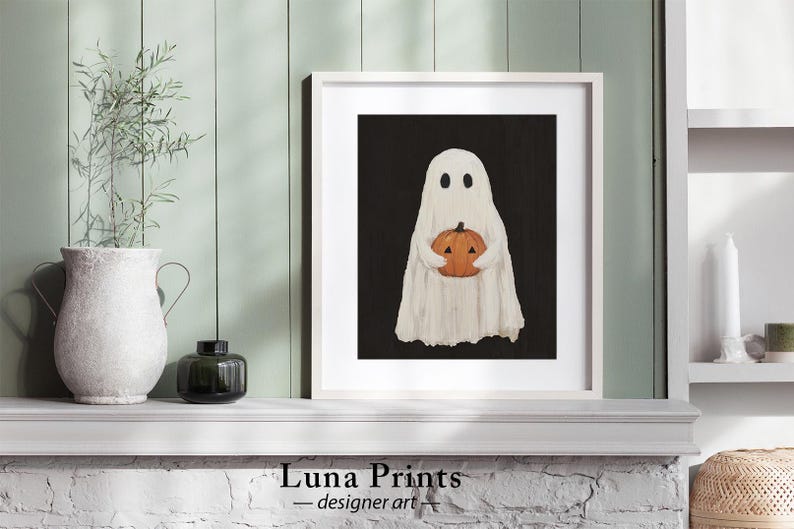 Peut inclure: Un fant&ocirc;me blanc tenant une petite citrouille orange devant un fond noir. Le fant&ocirc;me a deux yeux noirs et une petite bouche. L'image est encadr&eacute;e dans un cadre blanc.