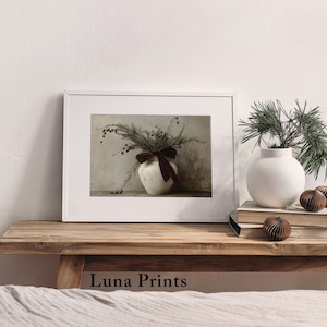 May include: A framed print of a white ceramic vase with a brown velvet bow, filled with dried foliage and berries. The vase is sitting on a wooden table with a white wall in the background.