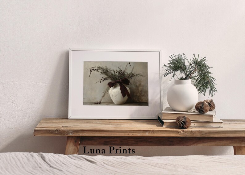 May include: A framed print of a white ceramic vase with a brown velvet bow, filled with dried foliage and berries. The vase is sitting on a wooden table with a white wall in the background.