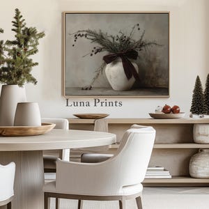 May include: A white ceramic vase with a burgundy ribbon sits on a table in front of a painting of a floral arrangement. The vase is surrounded by small potted trees and a wooden table with a bowl of fruit. The painting is titled "Luna Prints".