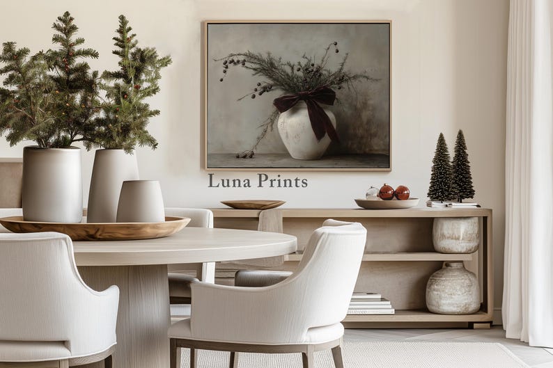 May include: A white ceramic vase with a burgundy ribbon sits on a table in front of a painting of a floral arrangement. The vase is surrounded by small potted trees and a wooden table with a bowl of fruit. The painting is titled "Luna Prints".