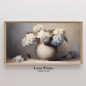 May include: A painting of a white vase filled with blue and white hydrangeas. The vase is sitting on a table with a light brown background. The painting is framed in a light wood frame.
