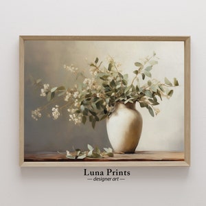 Puede incluir: Una pintura de un jarrón blanco lleno de flores blancas y hojas verdes. El jarrón está sobre una mesa de madera. El fondo es de color marrón claro. La pintura está enmarcada en un marco de madera claro. El texto "Luna Prints - designer art" está en la parte inferior de la pintura.