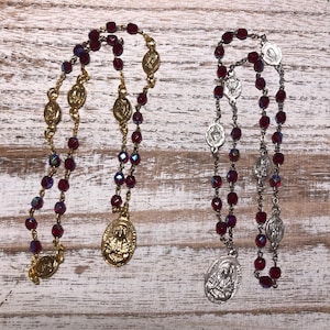 May include: Two rosaries with red beads and metallic accents. One rosary has gold-coloured metal components, including a crucifix and decorative medallions. The other rosary has silver-coloured metal components. Both rosaries are displayed on a wooden surface.