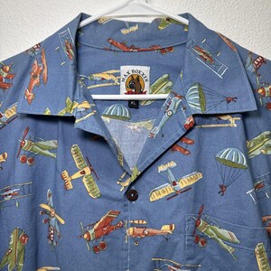 May include: Blue button-down shirt with an all-over print of vintage airplanes in red, yellow, green, and blue. The shirt has a collar and a single brown button. The label reads "MAX BOXXER" and "XL".