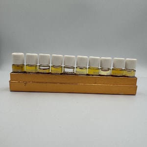 Op de afbeelding: Een set van tien kleine glazen parfummonsterflesjes met witte schroefdoppen, gerangschikt in een enkele rij in een rechthoekige goudkleurige kartonnen displaydoos. Elk flesje bevat een vloeistof en is voorzien van een etiket met tekst.