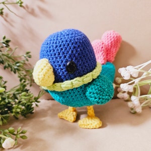 May include: A crocheted bird toy with a blue head, yellow beak, green body, and pink tail feathers.